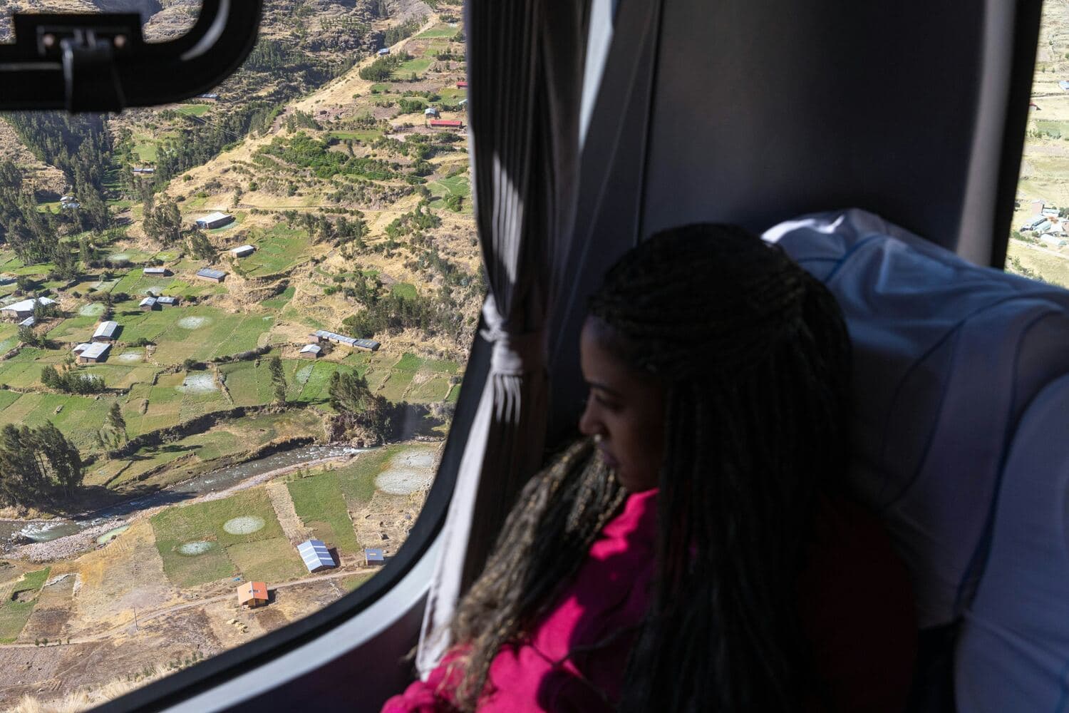 Una joven mira por la ventana, desde lo alto, un valle de montaña con campos y casas